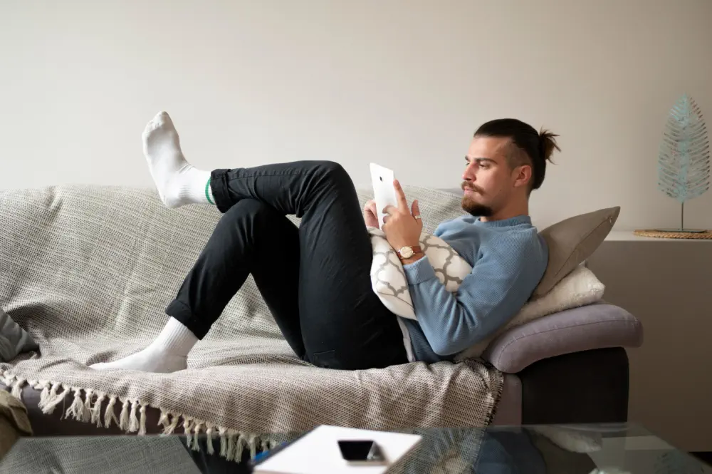 Man relaxes on a sofa, reading about alcohol withdrawal risks on his tablet.