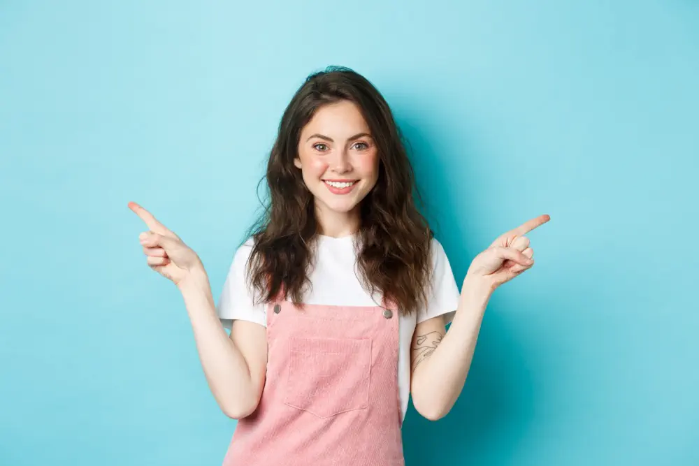 Smiling woman in pink overalls pointing sideways.